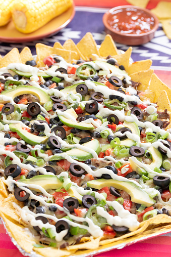Oven-baked sheet pan nachos layered with savory Mexican ingredients : tortilla chips, chicken, brisket, black beans, jalapenos, cheese, topping with avocados, green onions and olives and served with sour cream, salsa and corn cobs