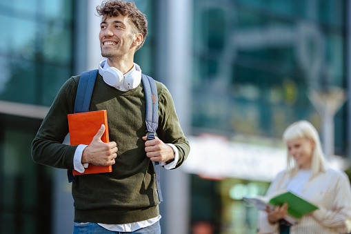 Cheerful male student with backpack standing on the street and holding a book