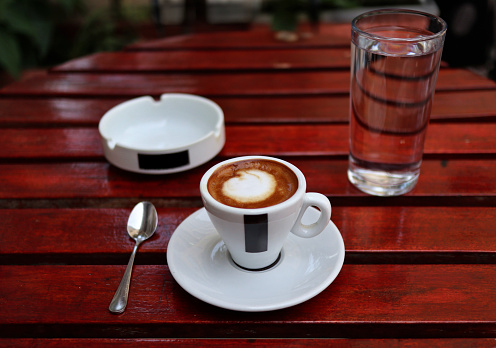 americano cup on saucer with spoon, ashtray, glass of water on red wooden coffee shop table (espresso, milk, macchiato) european style (cappuccino)