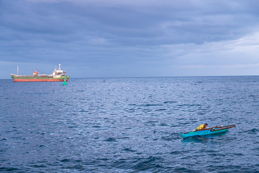 Dumaguete,Negros Island,Philippines-February 01 2023:At dusk, the man casts nets and dives in the water for fish and a large ship is anchored close beyond,at dusk.