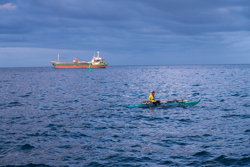 Dumaguete,Negros Island,Philippines-February 01 2023:At dusk, the man casts nets and dives in the water for fish and a large ship is anchored close beyond,at dusk.