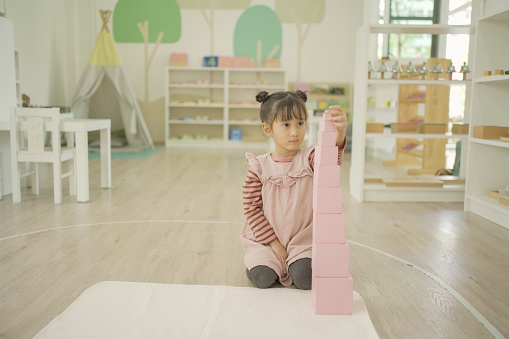 Preschool mixed girl building pink tower to learn a concept of size in three dimensions, first sensory material in Montessori education school