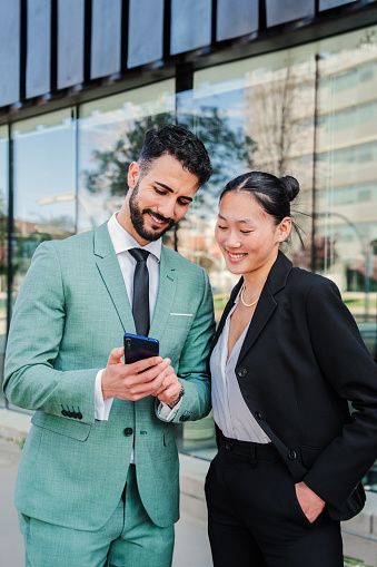 Vertical portrait of a couple of young businesspeople watching their successful stock shares on smartphone. Two partners smiling using a cellphone to invest on market. Coworkers with a mobile phone. High quality photo
