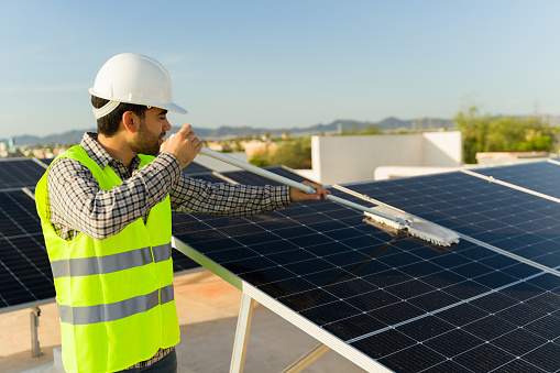 Latin man engineer with a vest and helmet cleaning the solar panels in the rooftop to clean energy generation