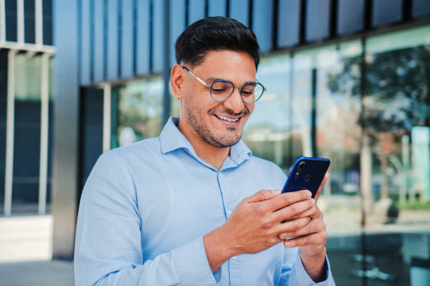 istock Hispanic young entrepreneur or business man smiling and using a cell phone app, wearing executive shirt and glasses. Handsome latin professional male browsing on internet with smartphone at workspace 1615217666
