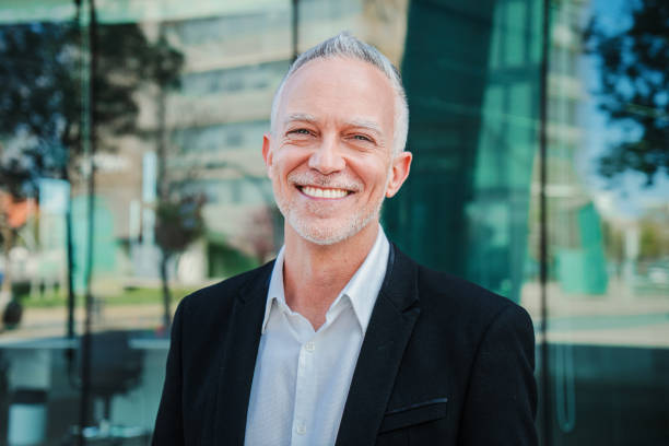 close up portrait of mature adult business man with gray hair and suit smiling and looking at camera with succesful attitude. happy corporate lawyer with white perfect teeth standing at workspace - old human face men ceo imagens e fotografias de stock