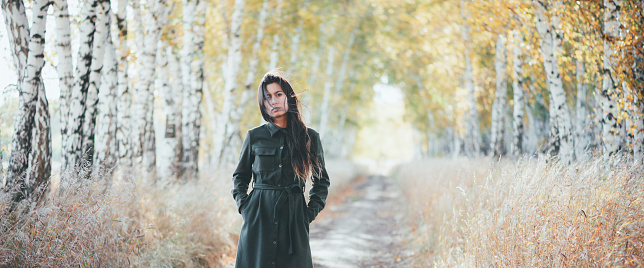 Sad beautiful girl with long natural black hair on bokeh background of autumn yellow leaves. Depressed girl in fall forest. Autumn apathy. Female emotional gloomy portrait in full growth. Faded tones.