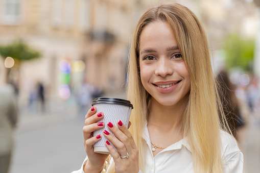 Happy blonde young woman enjoying morning coffee hot drink and smiling outdoors. Relaxing, taking a break. Teenager girl walking in urban city street, drinking coffee to go. Town lifestyles outside