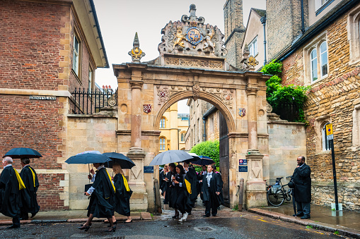 People exit Trinity College during Graduation Ceremony in Cambridge, Cambridgeshire, England, UK on a rainy day.