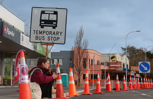 Tauranga New Zealand - July 31 2023; Road cones dominate street landscape while woman sits on kerb under temporary bus sign smoking and looking mobile device