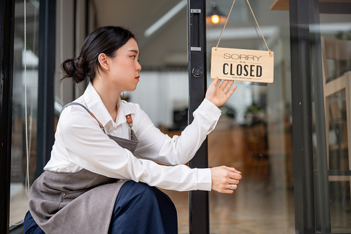 A sad and upset young Asian female coffee shop owner is hanging a close sign at the entrance door, feeling sad to close her business.