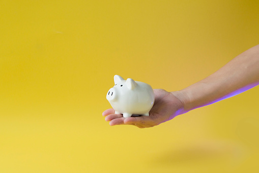 Hand holding white piggy bank on yellow background still life.