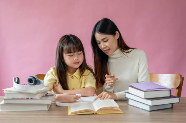 A kind Asian female teacher helps a young, adorable girl read a book and do homework A kind and caring Asian female teacher helps a young, adorable girl read a book and do homework at a study table against an isolated pink background. asian kids learning stock pictures, royalty-free photos & images