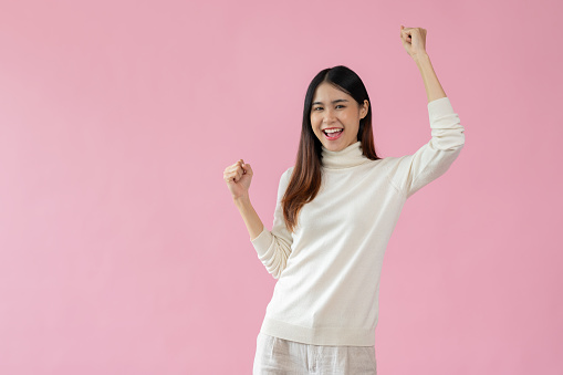 A cheerful and beautiful Asian woman is raising her hands, showing her fists, rejoicing, and standing against an isolated pink background with a joyful celebrating gesture.
