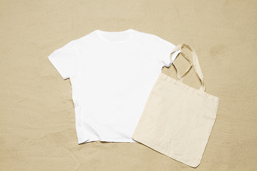 Mockup white not wrinkled summer t-shirt shopper copy space. Sand beach texture background. Blank template woman man shirt Top view. Summertime accessories. Flat-lay closeup tshirt. Beachtime