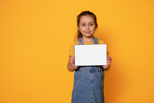 Adorable little child girl wearing a yellow stripped t-shirt and blue denim dress, showing a digital tablet with white empty blank screen, smiling looking at camera, isolated yellow studio background
