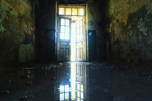 Open door in old abandoned shabby dark corridor with reflection in water. Exit to light from creepy haunted place.