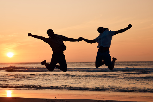 Beach sunset, shadow and silhouette of couple jump, holding hands and having fun, bonding and enjoy nature freedom. Sky, ocean sea and excited man, outdoor woman or people celebrate travel vacation