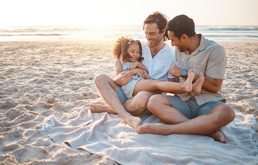 Gay parents, father and girl at beach with love, smile and hug for vacation, laugh or outdoor on sand in sunset. LGBTQ men, young kid and adoption with family, holiday or comic conversation in summer