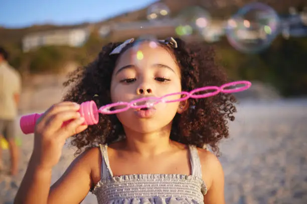 Photo of Girl child, beach and blowing bubbles with playing, outdoor and freedom on sand, games and thinking in nature. Female kid, soap and water with plastic toys with wind, summer sunshine and vacation