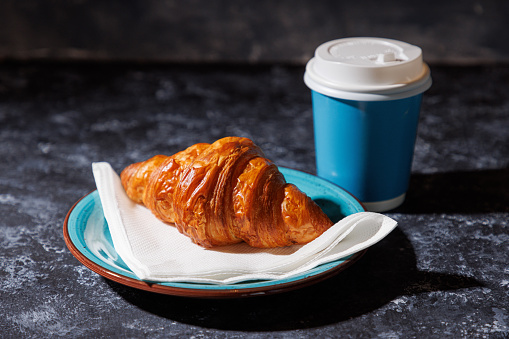 Beautifully baked crusty croissant on a plate in cafeteria, served with coffee in paper cup