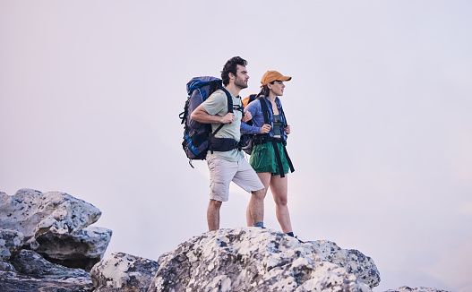 Mountains, hiking and man with woman on cliff for adventure in nature, landscape and travel. Outdoor trekking, couple on peak and relax in scenic clouds for natural journey, walking and rock climbing