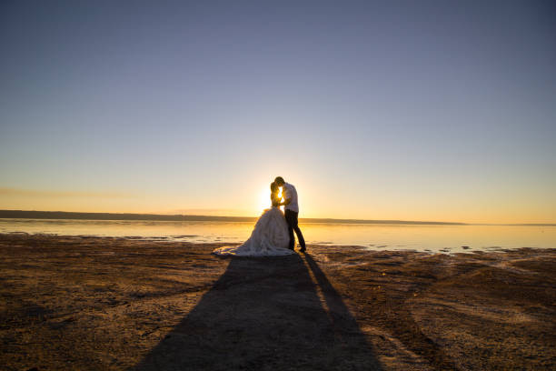 夕日を見ながら海のビーチでキスをする結婚式の夫婦。晴れた夏の写真。電車でオフショルダーのドレスを着て髪を下ろした花嫁。海のロマンチックな儀式。海辺の日の出のラブストーリー� - party beach indian ethnicity adult ストックフォトと画像