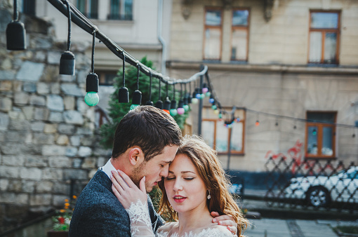 Wedding couple hugs in the old city. Rustic bride with hair down holds face of a groom in grey suit and bow tie. Love in ancient medieval town. White lace wedding dress. Vintage architecture details.