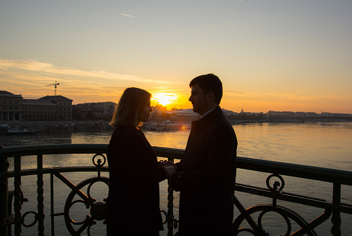 Couple in love is standing on the liberty bridge over the river Danube in Budapest. Sunrise in the big city. Dark silhouettes of man and woman holding hands in urban landscape. Sun kiss.