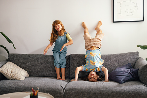 Young boy and girl playing on the sofa at home. The young girl is sitting on the sofa's backrest while the cute boy is upside down doing a handstand against the wall.