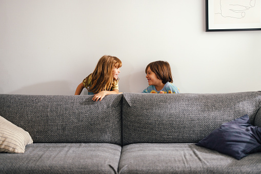 Portrait of two kids hiding behind the sofa at home. The young boy is smiling while looking at his sister. They are being playful.