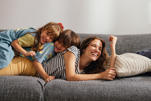 Portrait of a beautiful woman playing and having fun with her kids at home. She is lying on her belly on the sofa while her son and daughter are piling on top of her. They are all smiling.