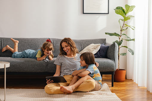Beautiful woman with her kids sitting on the floor and using digital tablet for playing games, watching movies and relaxing at home. Her son is cuddled with her, while the daughter is lying on the sofa next to them. They are all smiling and looking down at the screen.