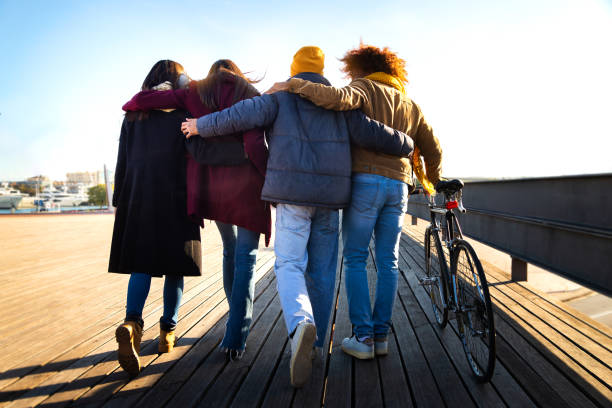 widok z tyłu grupy przyjaciół spacerujących razem, cieszących się słonecznym zimowym popołudniem. wielorasowi przyjaciele obejmujący się w mieście - port de barcelona zdjęcia i obrazy z banku zdjęć