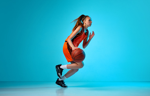 Young girl, basketball player in motion, running with ball against blue studio background in neon light. Concept of professional sport, action and motion, game, competition, hobby, ad