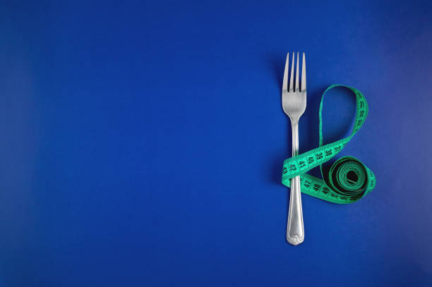 Measuring tape threaded through a fork on a blue background. Good proper nutrition. Medical fasting. The concept of a diet for weight loss. Healthy lifestyle.