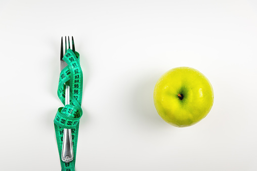 A metal fork wrapped with measuring tape next to a green apple on a white background. The concept of losing weight and losing excess weight and fat.