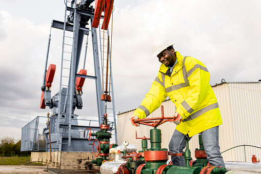 Oil field worker closing pipeline valve. In background oil rig producing crude oil.