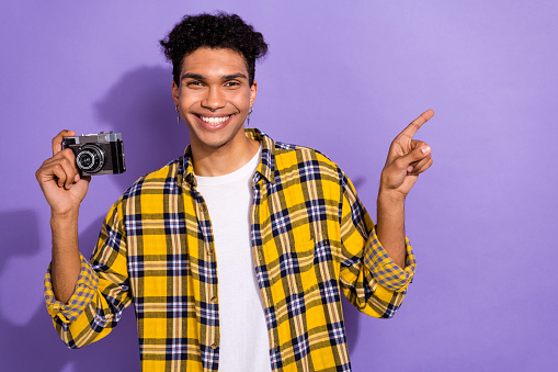Photo of cheerful attractive guy wear plaid yellow shirt rising vintage camera pointing empty space isolated violet color background.