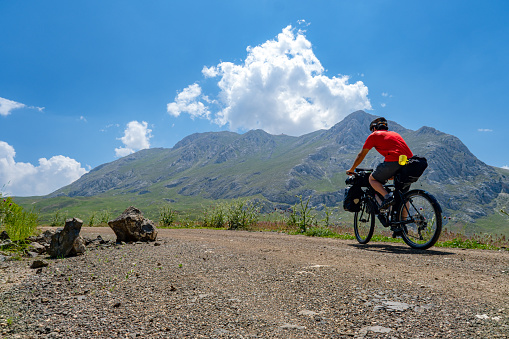 Tour bike in bikepacking order. Cyclist in red jersey with helmet. High altitude mountainous area Mediterranean region. Dirt road. Camper riding bike. Side angle. Less cloudy weather clear sunny. Steep mountainous region.