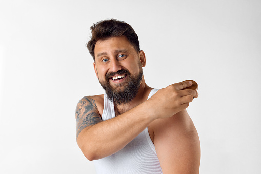 Delightful, smiling, bearded man in singlet taking care after appearance at home over white studio background. Concept of men's beauty, skin care, cosmetology, health and wellness. Copy space for ad