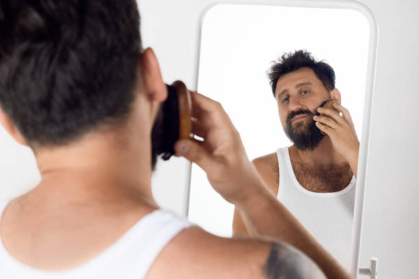 istock Mature man looking in mirror, taking care after beard, brushing with special brush against white studio background. 1616148897