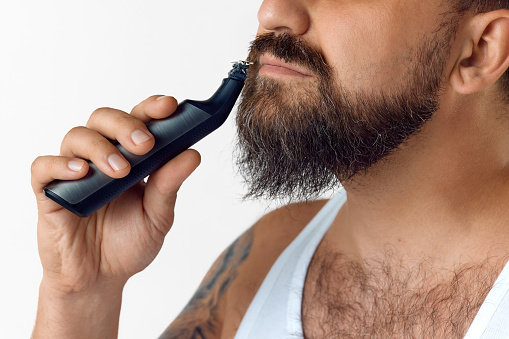 Cropped image of man taking care after beard with shaver against white studio background. Concept of men's beauty, skin care, cosmetology, health and wellness. Copy space for ad