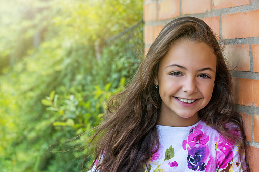 Little smiling long haired girl is posing in the garden leaning against a brick wall. Happy childhood concept. Horizontally.