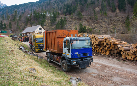 Forni Avoltri, Italy - April 8th 2023. A flatbed truck loaded with timber and a container truck at a saw mill yard near the village of Forni Avoltri in Carnia, Udine Province, Friuli-Venezia Giulia, north east Italy. The building is a former restaurant