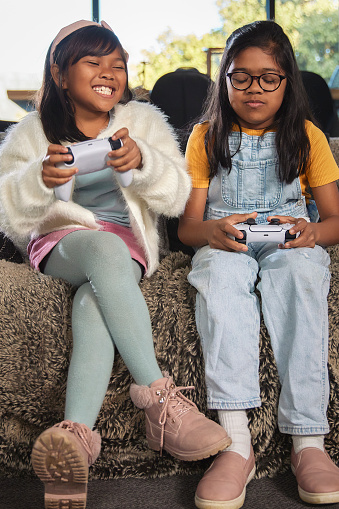 In this lively portrait, two sisters are engrossed in a video game. One sister is visibly excited, her animated expressions reflecting her joy in the game. The other sister wears a slightly grumpy expression, likely from losing the game. The contrast in their emotions adds a touch of realism to the scene, capturing the dynamic range of emotions that can arise during friendly competition and shared activities.