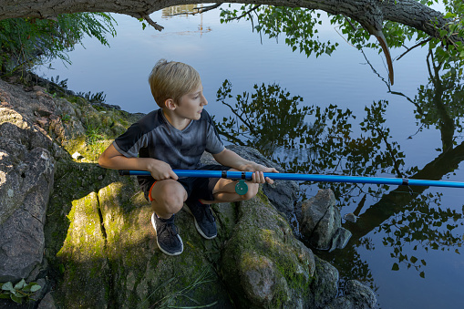 A boy squats on rocks and catches fish. Sport fishing on the river in summer.