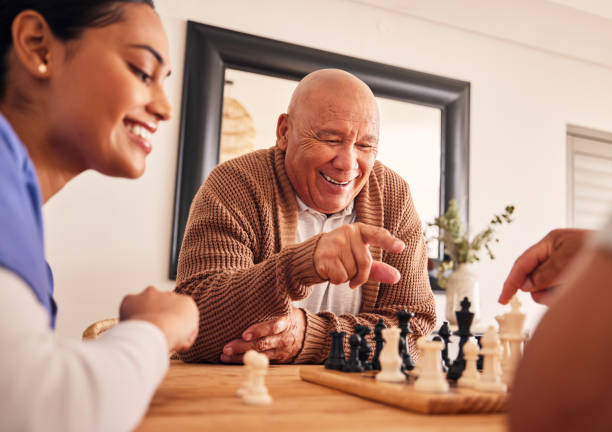 senior man, nursing home and chess for game, competition and happy with friends, strategy and relax together. elderly person, nurse and board for contest with mindset, excited smile or point at table - ouderenzorg stockfoto's en -beelden