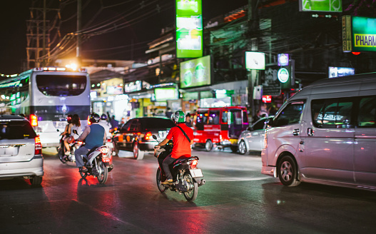A busy street at night in Phuket. The main feature of the image is the motorbikes that are in the foreground, with one rider wearing a red shirt and helmet. The background shows buildings with lit up signs, including a pharmacy and a green sign with white text. The street is lined with power lines and street lights, creating a chaotic and bustling atmosphere.