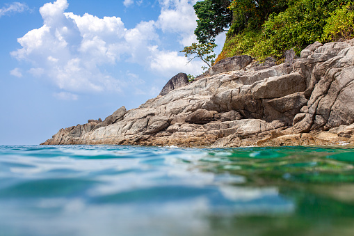 A gray and jagged rocky coastline with green trees and bushes on top. A bright blue water that is calm with small waves, reflecting the sky. A clear blue sky with a few white clouds, creating a contrast between the light and dark colors. A perspective from the water, looking up at the rocks and sky, as if the viewer is swimming or kayaking. A photo realistic image of a natural landscape.